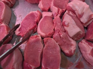Raw pork loin chops or  loin cuts with white fat edges on a tray, shown with metal tongs under bright lighting, ideal for butchery or food handling imagery.