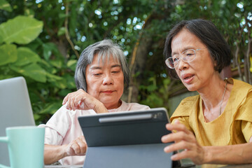 Two senior Asian women friends learning and using a tablet computer together outdoors. Elderly lifestyle with modern technology and online connection concept.