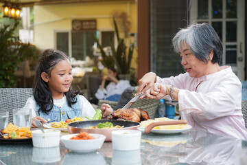 Happy Asian grandmother and granddaughter enjoying a family meal together at an outdoor restaurant. The senior woman cuts chicken for the young girl. Intergenerational bonding concept.