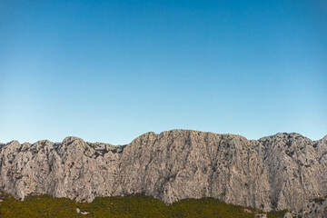 A panoramic view of a rocky mountain range under a clear blue sky. The landscape features rugged cliffs and green vegetation at the base.