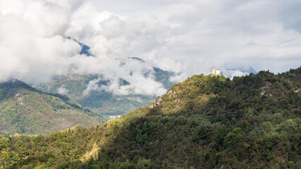 Scenic view of San Floriano Basilica on the hill of Illegio, Friuli Venezia Giulia, surrounded by lush green forests and dramatic mountain clouds.