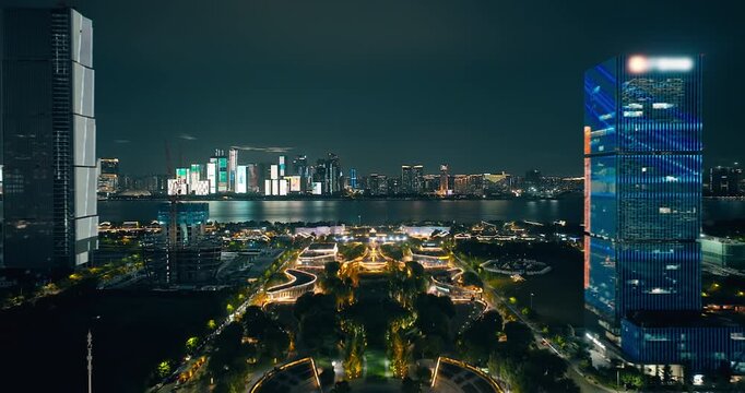 Aerial shot of a futuristic city skyline illuminated across a river at night with a modern park in the foreground. Urban nightscape background.