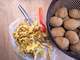 Preparing potatoes in the kitchen – unpeeled potatoes in a plastic basket, potato peels, knife and peeler on a wooden table. Homemade cooking preparation scene.
