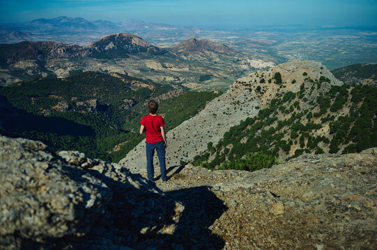 A young hiker stands on a rocky ridge, gazing over rolling hills and distant mountains. The red shirt pops against rugged terrain, capturing exploration and awe.