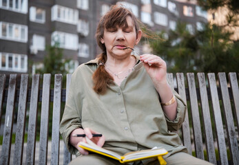 Elderly woman draws in notebook, pursues hobby, sitting on bench in city park. Active age, interesting life and creative abilities of older people