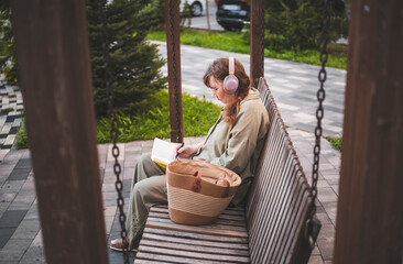 Aged woman in headphones sits thoughtfully drawing in notebook, pursues hobby, in city park. Active age, interesting life and creative abilities of older people