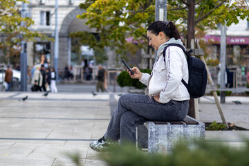Obraz premium A girl with black hair in a white jacket in a park on a cloudy day looking at her smartphone