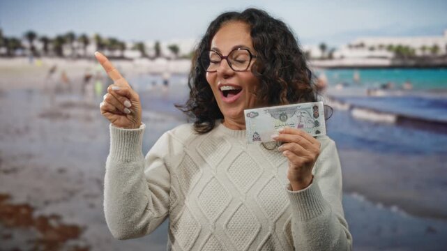 Middle-aged woman at seaside in uae holds a dirham banknote looking surprised and pointing, with a beach and water background showcasing a lively outdoor scene.