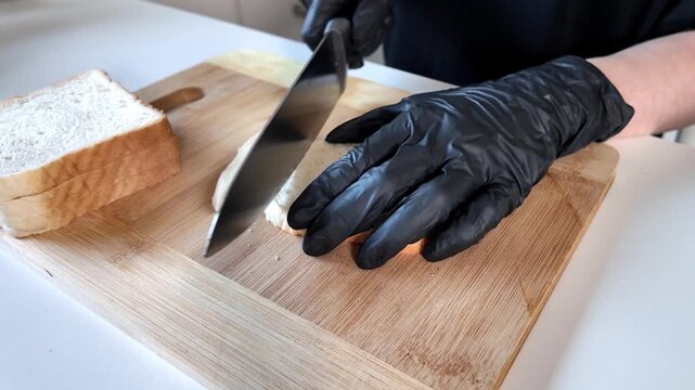 Skilled gloved hands carefully slice fresh white bread on a rustic wooden cutting board, emphasizing hygienic food preparation and professional culinary practice