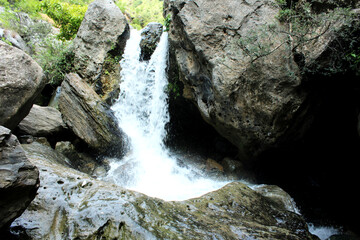 A small, clear waterfall cascading over and through large, sunlit gray boulders and rocks in a rocky gorge, surrounded by dense green foliage