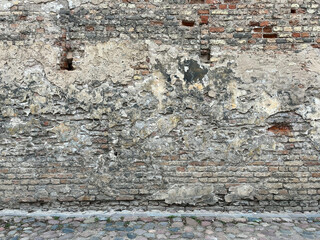 Weathered brick wall with deteriorated plaster, cracks, and holes. Cobblestone ground adds rustic charm. Ideal for urban decay, texture, or historical design themes.