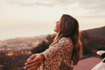 Young beautiful woman enjoying summer and having fun sitting on the hood of the car opening arms feeling good and free. Freedom concept and happy lifestyle at sunset