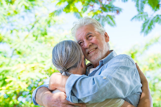 Beautiful cute couple of seniors hugged having fun and enjoying together outdoors in green park smiling looking around them. Two pensioners embracing taking care of relationship