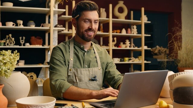Happy joyful smiling Caucasian man small business owner craftsman businessman potter crock male working laptop typing desktop browsing computer online shopping pottery selling inside workshop studio - Powered by Adobe