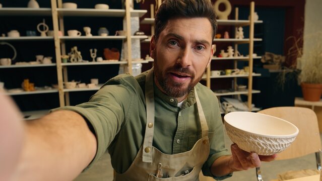 Talented Caucasian salesman potter male man guy businessman artist small business owner holding pottery ceramic bowl record video blog on mobile phone talking looking webcam camera in workshop store