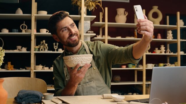 Happy smiling Caucasian man male guy small business owner potter entrepreneur blogger artist using mobile phone record video online blog social media showing pottery bowl photo selfie indoor workshop
