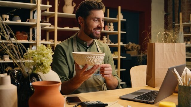 Happy joyful creative Caucasian talented craftsman man male guy businessman artisan small business owner holding pottery bowl decoration stoneware talking online video call laptop computer sell indoor