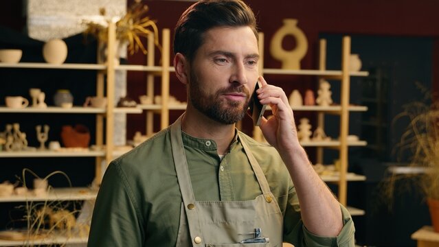 Professional skilled potter Caucasian man male guy businessman small business owner seller potter ceramist artist craftsman talking mobile phone deal call client discussing order in pottery workshop