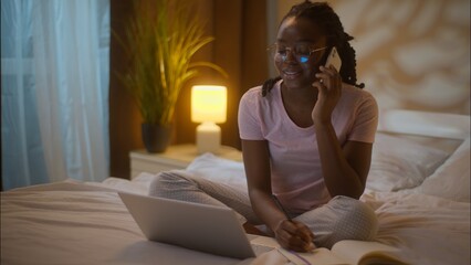 Multitasking African American business woman student female girl businesswoman in glasses talk mobile phone call working laptop freelance typing computer writing notes sitting bed at home late evening