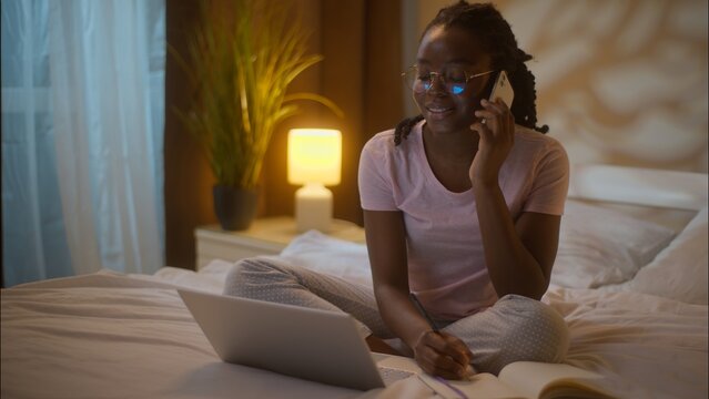 Multitasking African American business woman student female girl businesswoman in glasses talk mobile phone call working laptop freelance typing computer writing notes sitting bed at home late evening