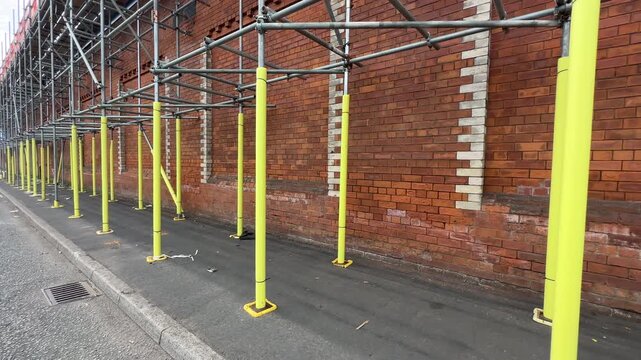 Personal POV walking in the street by long scaffolding erected for renovation of an old typical red brick building in Salford city.
