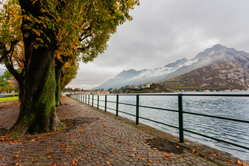 Late-afternoon view of Lake Como from Lecco, Italy, during autumn. Vibrant red, orange and green trees line the calm lakeshore beneath dramatic grey clouds. A distant couple stands by the water as the