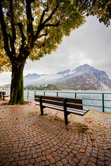 Late-afternoon view of Lake Como from Lecco, Italy, during autumn. Vibrant red, orange and green trees line the calm lakeshore beneath dramatic grey clouds. A distant couple stands by the water as the