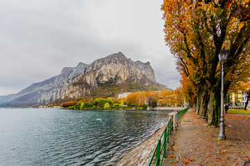 Late-afternoon view of Lake Como from Lecco, Italy, during autumn. Vibrant red, orange and green trees line the calm lakeshore beneath dramatic grey clouds. A distant couple stands by the water as the
