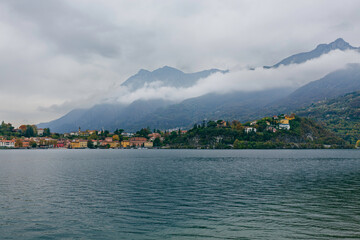 Late-afternoon view of Lake Como from Lecco, Italy, during autumn. Vibrant red, orange and green trees line the calm lakeshore beneath dramatic grey clouds. A distant couple stands by the water as the