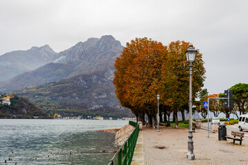 Late-afternoon view of Lake Como from Lecco, Italy, during autumn. Vibrant red, orange and green trees line the calm lakeshore beneath dramatic grey clouds. A distant couple stands by the water as the