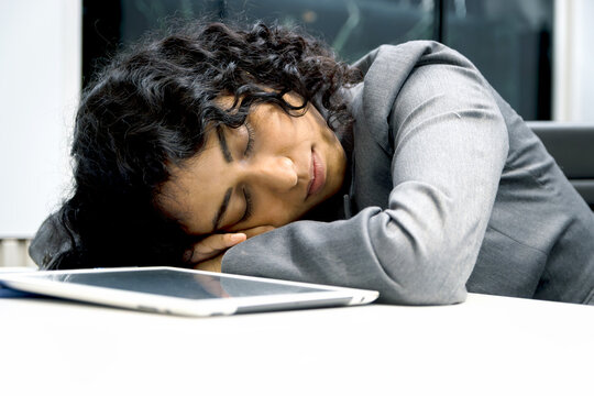 Exhausted young businesswoman with curly hair working hard overtime, sleeping in office desk. Tired beautiful staff officer feels sleepy after overwork project, need to take a nap at workplace.