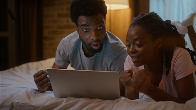 Happy excited African American woman girlfriend boyfriend man lying on bed family couple watching digital tablet shocked sport match game victory celebrating win shouting triumph at home bedroom night