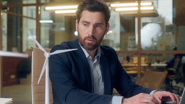 Depressed Caucasian businessman office engineer looking wind turbine model eco startup struggles sustainability doubts green energy project challenge laptop thinking plan innovation environment future - Powered by Adobe