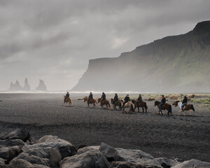 Group of horseback riders riding along a black sand beach in Iceland, surrounded by dramatic cliffs and misty coastal scenery. The moody atmosphere and natural setting create a cinematic, adventurous 