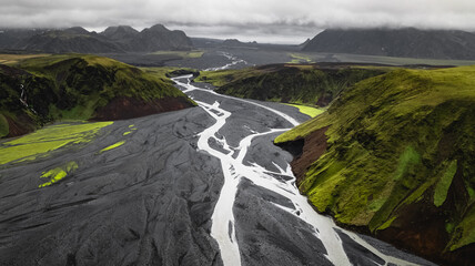 Aerial view of a braided river flowing through a dramatic volcanic landscape in the Icelandic highlands. The contrast between black volcanic sand, vibrant green moss-covered hills, and winding white g