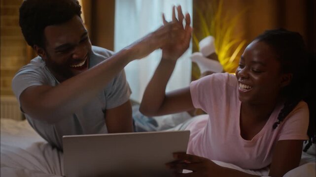 Overjoyed happy African American family ethnic couple woman man lying bed watching digital tablet online sport match betting giving high five gesture support victory winning luck success home bedroom - Powered by Adobe