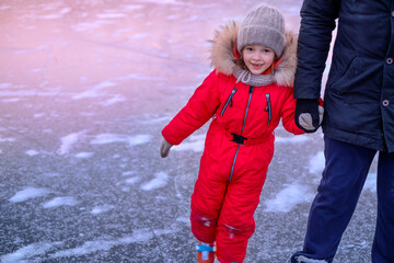 Funny laughing little girl in a red suit skating on a frozen lake holding her dad's hand