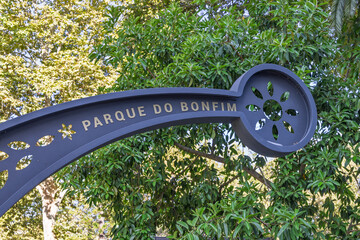 Close-up of the ornate, dark metal sign for Parque do Bonfim in Setúbal, featuring gold lettering and a decorative wheel-like motif against lush greenery.