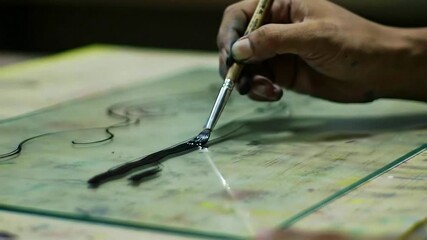 A close-up video shows an artist's steady hand skillfully painting a thick black line onto a sheet of glass demonstrating traditional craftsmanship and artistry.
