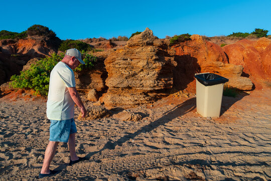 Hombre mayor caminando hacia la basura en la playa, un d&iacute;a soleado con cielo azul..