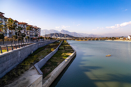 Scenic river view from bridge showing Boğa Çayı with waterfront promenade, residential buildings, and hazy Taurus Mountains under winter sun. Picturesque city corner in Antalya, Turkey.
- Powered by Adobe