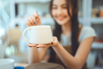 Close-up of handmade ceramic cup in pottery studio with happy Asian woman thinking of creative...