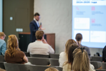 Speaker at podium presents to group of professionals in bright modern conference room with slides projected on screen behind