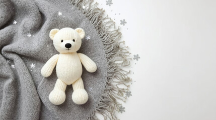 A charming hand-knit bear toy rests on a gray woven throw with fringe details, scattered with decorative snowflake ornaments. The scene evokes feelings of warmth, comfort, and cozy wintertime charm.