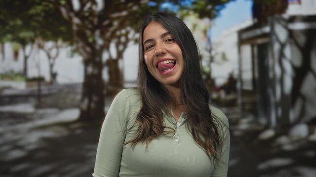 Young hispanic woman in green top sticks tongue out on sunny street framed by dappled tree shadows; playfulness.