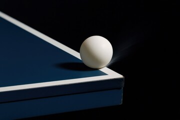 A white ping pong ball is perched on the edge of a blue table, with a slight motion blur indicating it is about to drop off the edge. The background is black, creating a high contrast effect.