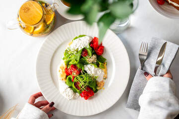 Top view of a fresh gourmet meal with green pasta, cherry tomatoes, cheese, and microgreens on a white plate. Elegant table setting with tea and hands ready to eat.
