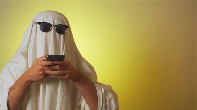 Halloween. Ghosting. Ghost costume with white blanket and sunglasses texting on smartphone over yellow background