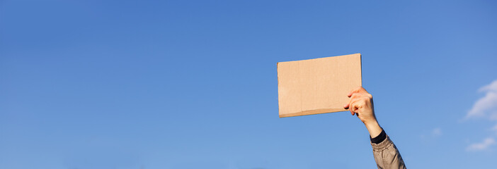Close-up of a hand raised up and holding a blank piece of cardboard for writing against a sky background. Protest or warning. The blank space, can be used to add text or a symbol.
