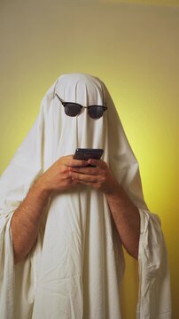 Halloween. Ghosting. Ghost costume with white blanket and sunglasses texting on smartphone over yellow background